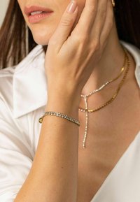 Goldarmband mit klaren Edelsteinen, glatte Textur, getragen an einem schlanken Handgelenk. Begleitet von einer geschichteten Kettenhalskette aus Gold und Silber.