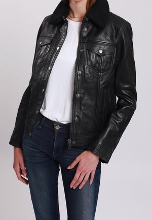 Nainen pukeutunut mustaan nahkatakkiin, jossa on napilliset rintataskut ja musta karvareunus, valkoisen t-paidan ja sinisten farkkujen päällä.