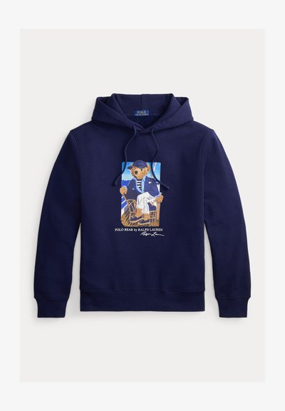 Μπλε σκουρόχρωμο hoodie με κορδόνι, με γραφικό ενός αρκούδου ντυμένου ναύτη που κάθεται σε καρέκλα και κείμενο "Polo Bear by Ralph Lauren" από κάτω.