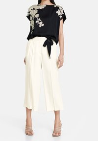 Zwarte zijden blouse met bloemmotief, losse fit, gestrikt op de taille. Crèmekleurige, wijde broek met een gladde textuur en plooien aan de voorkant. Neutrale sandalen met banden.