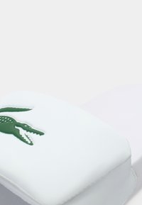 Sandale slide blanche avec une tige rembourrée et un logo crocodile vert. Texture lisse et forme arrondie avec des bords cousus.