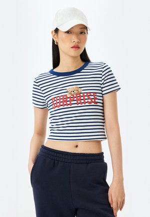 Ingekorte marineblauwe en witte gestreepte t-shirt met rode tekst "SURPRISE" en een teddybeer ontwerp. Draagt het met donkere broek en een witte pet.