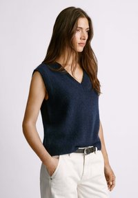 Navy blue bezrukávový sveter s výstrihom do V, vyrobený z mäkkej pleteniny, s rebrovaným lemom a uvoľneným strihom. Nosený cez svetlé nohavice.