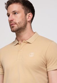 Homme aux cheveux bruns courts et à la barbe, portant un polo beige avec deux boutons et un petit logo brodé sur la poitrine.
