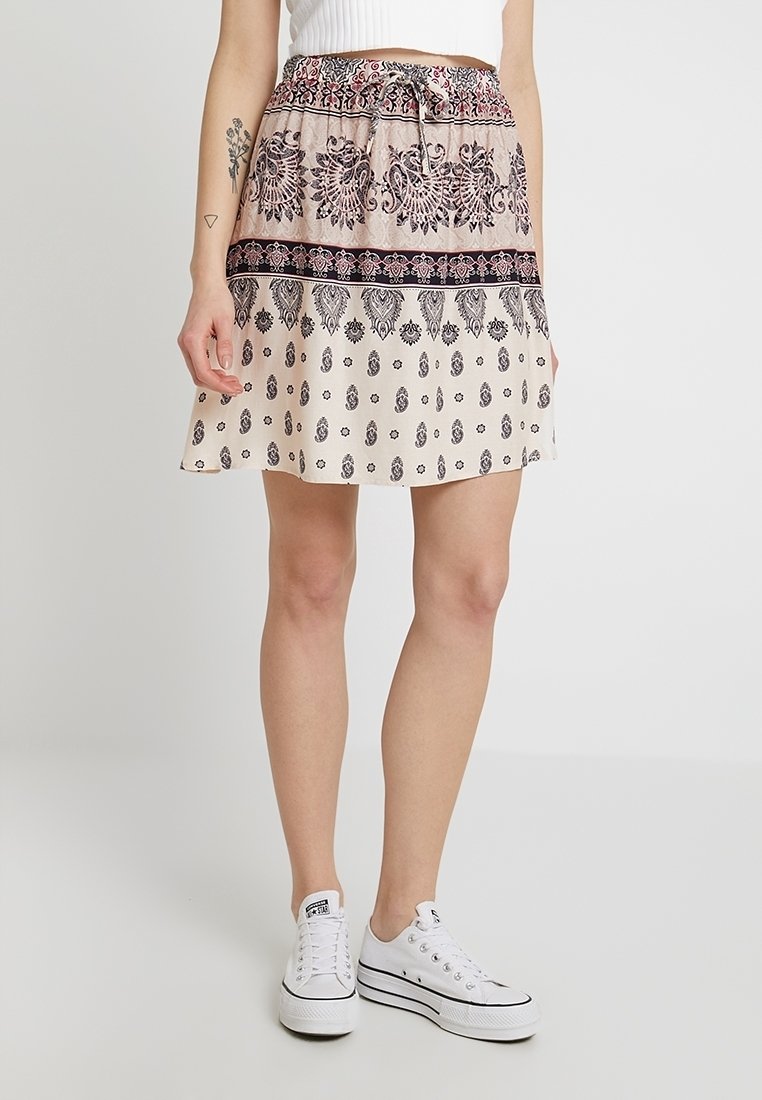 Lyhyt, laventuva hame beige-värissä, jossa on monimutkaisia mustia kuvioita ja nauhavyötärö. Yhdistetty valkoisiin lenkkareihin rentoa ilmettä varten.