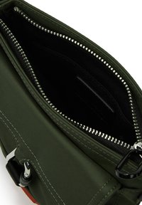 Πράσινη νάιλον τσάντα με μαύρη επένδυση, λεπτομέρεια από ασημένιο φερμουάρ και ορθογώνια ετικέτα. Υφασμάτινη επιφάνεια με ορατά ράμματα.