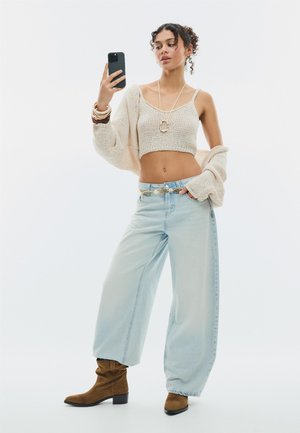 Mladá žena s kučeravými vlasmi, ktorá nosí béžový crop top, kardigán, široké rifle a hnedé čižmy, a robí si selfie čiernym smartfónom.