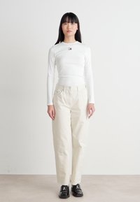 Hvit langermet topp med en liten logo på brystet, kombinert med løse beige bukser og svarte snørebåndssko.