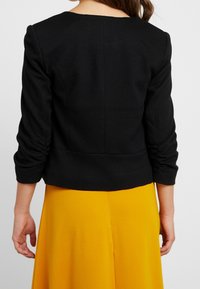 Black textured jacket with puffed sleeves, fitted waist, and rounded neckline, paired with a vibrant yellow skirt.