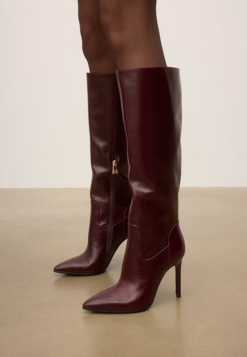 Bottes en cuir marron foncé montant jusqu'aux genoux, à bouts pointus, talons hauts et fins, et fermetures éclair latérales, portées sur jambes nues.