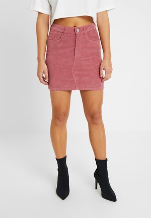 Vrouw die een roze corduroy minirok, een wit cropped topje en zwarte, puntige, hoge enkellaarsjes draagt, staand op een witte vloer.