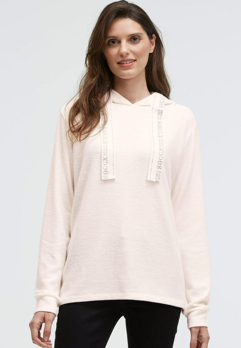 Young woman with long brown hair wearing a loose white hoodie with printed drawstrings and black pants, standing against a plain background.