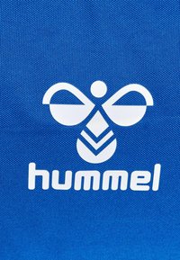 Fondo de tela azul con un logo blanco que presenta un diseño abstracto de abeja sobre el texto "hummel" en una fuente negrita y redondeada.