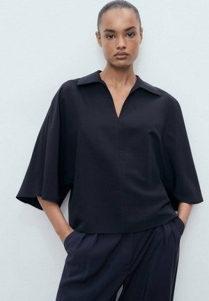 Túnica negra de gran tamaño con un amplio cuello, escote en V y mangas cortas y anchas. La tela es suave y tiene un diseño minimalista, combinada con pantalones oscuros.