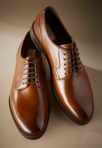 Zapatos de vestir de cuero marrón con una superficie lisa y brillante, puntera redonda y seis ojetes para los cordones. Detalles de costura añaden textura.