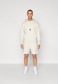 Completo da tuta Nike beige chiaro composto da felpa con cappuccio e stampa del logo, abbinato a pantaloncini, entrambi realizzati in tessuto morbido. Inclusi scarpe bianche.