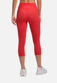 Leggings capri rosse a vita alta con una texture liscia, caratterizzate da dettagli cuciti bianchi e marchi etichettati sulle gambe inferiori. Indossati con sneaker bianche.