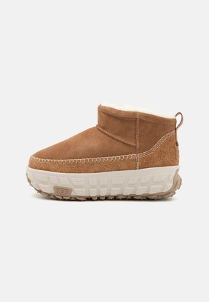 Brązowe zamszowe botki za kostkę, z miękką, białą futrzaną wyściółką oraz teksturowaną gumową podeszwą z wyraźnymi wzorami bieżnika.