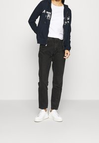 Marineblauwe zip-up hoodie met voorzakken, voorzien van een logo-afdruk. Gecombineerd met high-waisted zwarte jeans en witte sneakers.