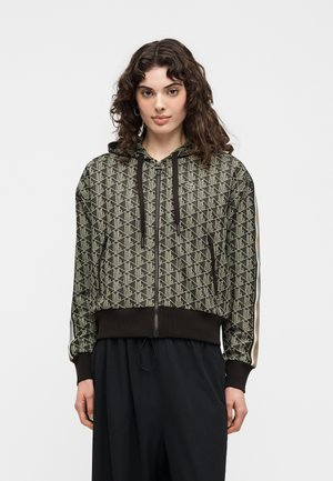 Femme aux cheveux bouclés portant un sweat à capuche zippé à motif géométrique avec des poignets noirs et des cordons de serrage, debout devant un fond uni.