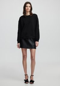 Sweatshirt noir avec un détail de lacets texturé sur les manches, associé à une jupe en cuir noir ajustée. Le modèle porte des talons aiguilles noirs.