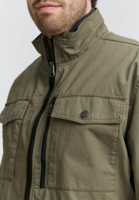 Olivegrüne Jacke aus leichtem Stoff mit hohem Kragen, durchgehendem Reißverschluss und Brusttaschen mit Druckknöpfen. Strukturiertes Finish.