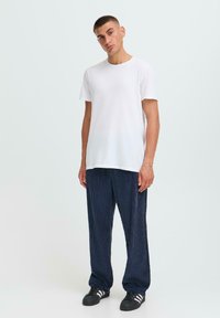 Un jeune homme se tient debout, portant un t-shirt blanc uni, un pantalon en velours côtelé bleu foncé ample et des baskets noires avec des rayures blanches.
