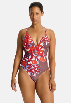 Woman wearing a one-piece swimsuit with red, white, and blue floral patterns on a brown background, standing against a plain white background.