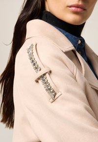 Lichtroze, zachte stoffen jas met een decoratieve kristallen embellissement langs de schouder. Klassieke kraag en knoopdetails zichtbaar.