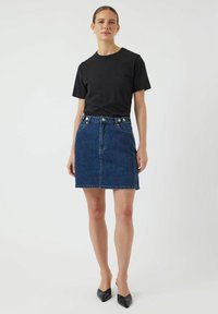 Hame denim berwarna biru gelap dengan siluet pas, desain lima kantong, dan penutupan kancing. Dipadukan dengan kaos hitam lengan pendek.