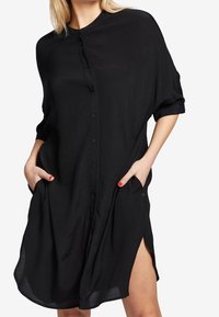 Svart skjortklänning i lätt material, med knäppning fram, korta ärmar, sidofickor och sidslitsar för bättre rörlighet.
