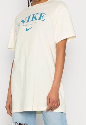 Osoba nosící volné krémové tričko Nike s modrým logem a textem, v kombinaci se světle modrými roztrhanými džínami.