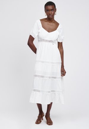 Frau steht und trägt ein weißes Midikleid mit Puffärmeln und Spitzendetails, kombiniert mit braunen Sandalen mit Absatz und Creolen.