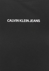 Texte blanc indiquant "CALVIN KLEIN JEANS", centré sur un fond en tissu noir uni.