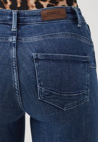 Tamno plave traperice s klasičnim dizajnom od pet džepova i smešnim kožnim oznakom s "ONLY" brendingom na stražnjem pojasu.