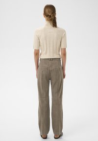 Vrouw met bruin paardenstaartje, gekleed in een beige, geribbelde korte-mouwen coltrui en losse taupekleurige broek, staande tegen een witte achtergrond, achterkant zichtbaar.