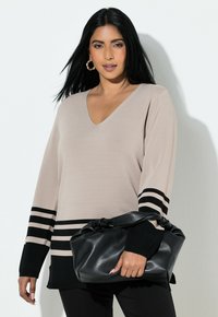 Béžový svetřík s výstřihem do V s černými vodorovnými pruhy na manžetách a lemu. Modelka drží černou koženou tašku s uzlovým uchycením.