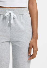 Graue Jogginghose mit weicher Textur, ausgestattet mit einem weißen Kordelzug mit aufgedrucktem Logo. Nahaufnahme des Taillenbunds und einer Hand, die den Stoff hält.