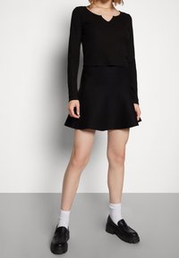 Top de manga larga negro con textura acanalada y escote con ondas, combinado con una falda negra acampanada. La modelo lleva mocasines negros gruesos y calcetines blancos.