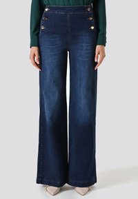 Jean large taille haute bleu foncé avec des accents de boutons dorés sur les poches et une texture en denim. Finition lisse avec une coupe ajustée.