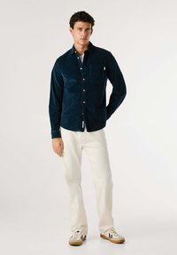 Marineblå kordfløyelsbutton-up skjorte med en brystlomme, kombinert med lyse bukser og sneakers. Glatt tekstur og slim fit-design.