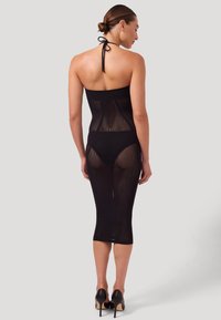 Donna in piedi, indossa un abito nero a rete con collo ad anello sopra biancheria intima nera e tacchi alti neri, di spalle su sfondo semplice.