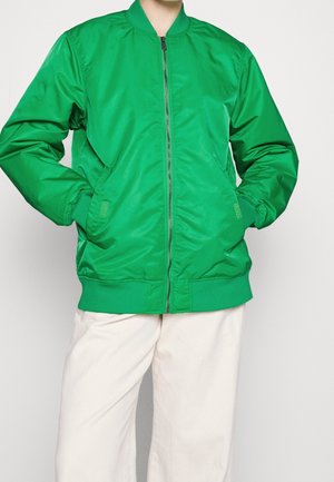 Henkilö, jolla on kirkkaanvihreä vetoketjullinen bomber-takki, kädet taskuissa ja väljät, luonnonvalkoiset housut yksinkertaisella taustalla.