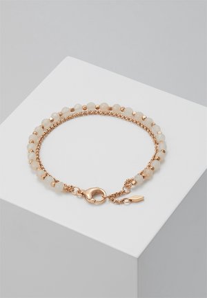 Gouden kettingarmband met kleine ronde bleke kralen en een karabijnsluiting, tentoongesteld op een witte schuinstaande plaat.