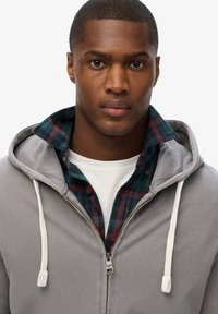 Gray zip-up hoodie with white drawstrings, layered over a green and red plaid shirt, and a white crew neck T-shirt underneath.