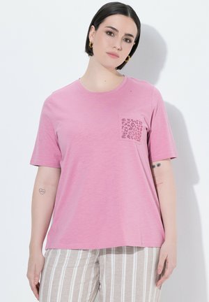 Vrouw met kort donker haar draagt een roze t-shirt met een patroon op de zak en gestreepte beige broek, staand tegen een witte muur.