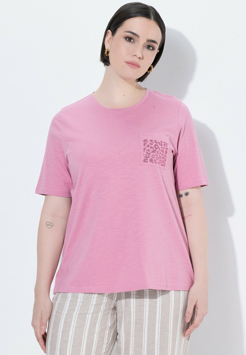 Femme aux cheveux courts foncés portant un t-shirt rose avec une poche à motif et un pantalon beige rayé, debout devant un mur blanc.