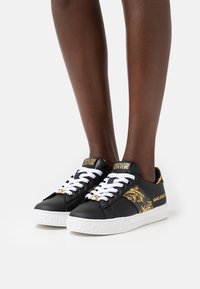 Feet wearing black sneakers with white laces, gold baroque pattern accents, and "Versace Jeans Couture" branding on tongue, sides, and sole.