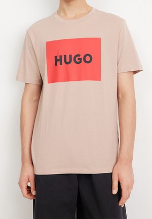 Jasnobeżowa bawełniana koszulka z wyraźnym czerwonym prostokątem, na którym znajduje się napis „HUGO” pogrubionymi czarnymi literami. Krótkie rękawy i okrągły dekolt.