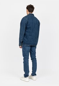 Donkerblauwe denimjack en spijkerbroek met een relaxte pasvorm en een klassieke button-down kraag. Witte sneakers maken de outfit compleet.